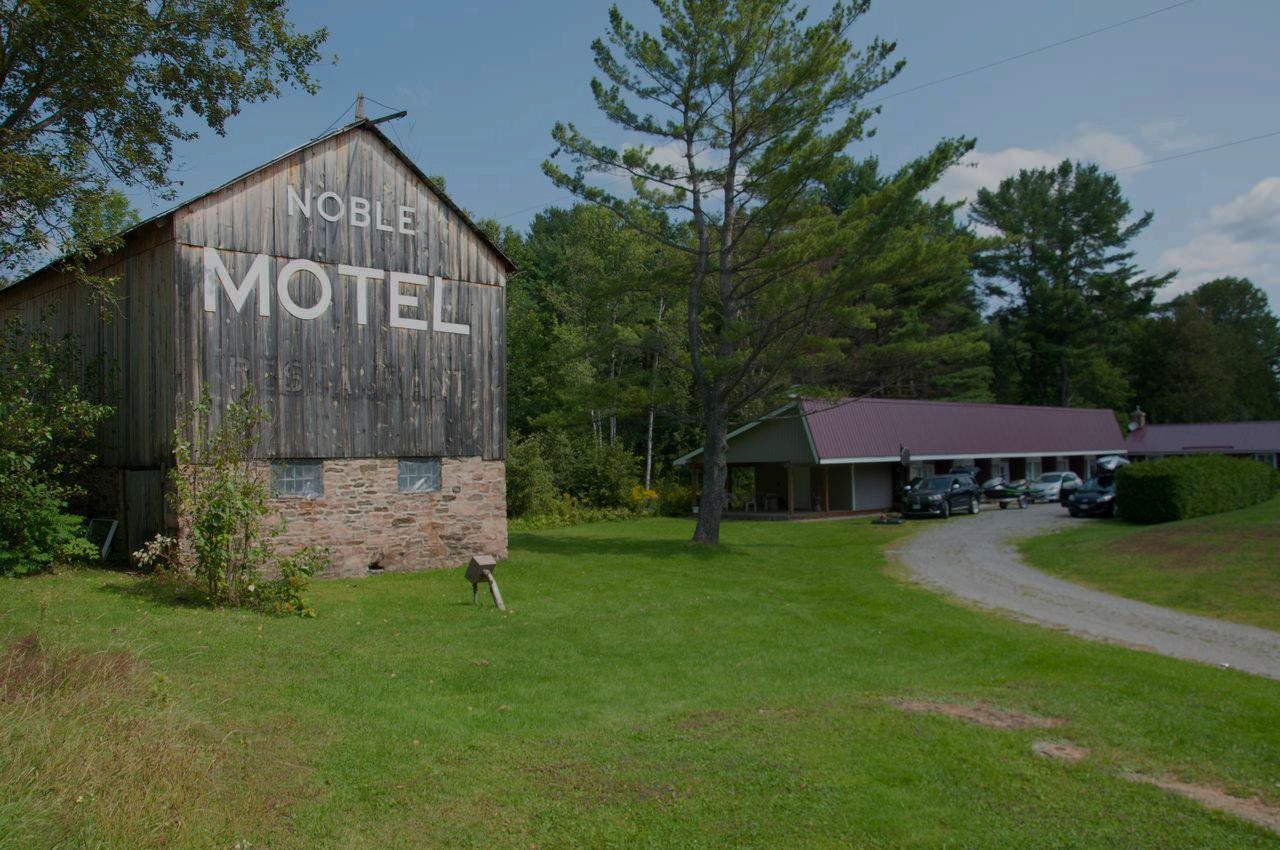 The Noble Motel's weathered barn with MOTEL painted across it, the guest room block and driveway behind, framed by tall pines